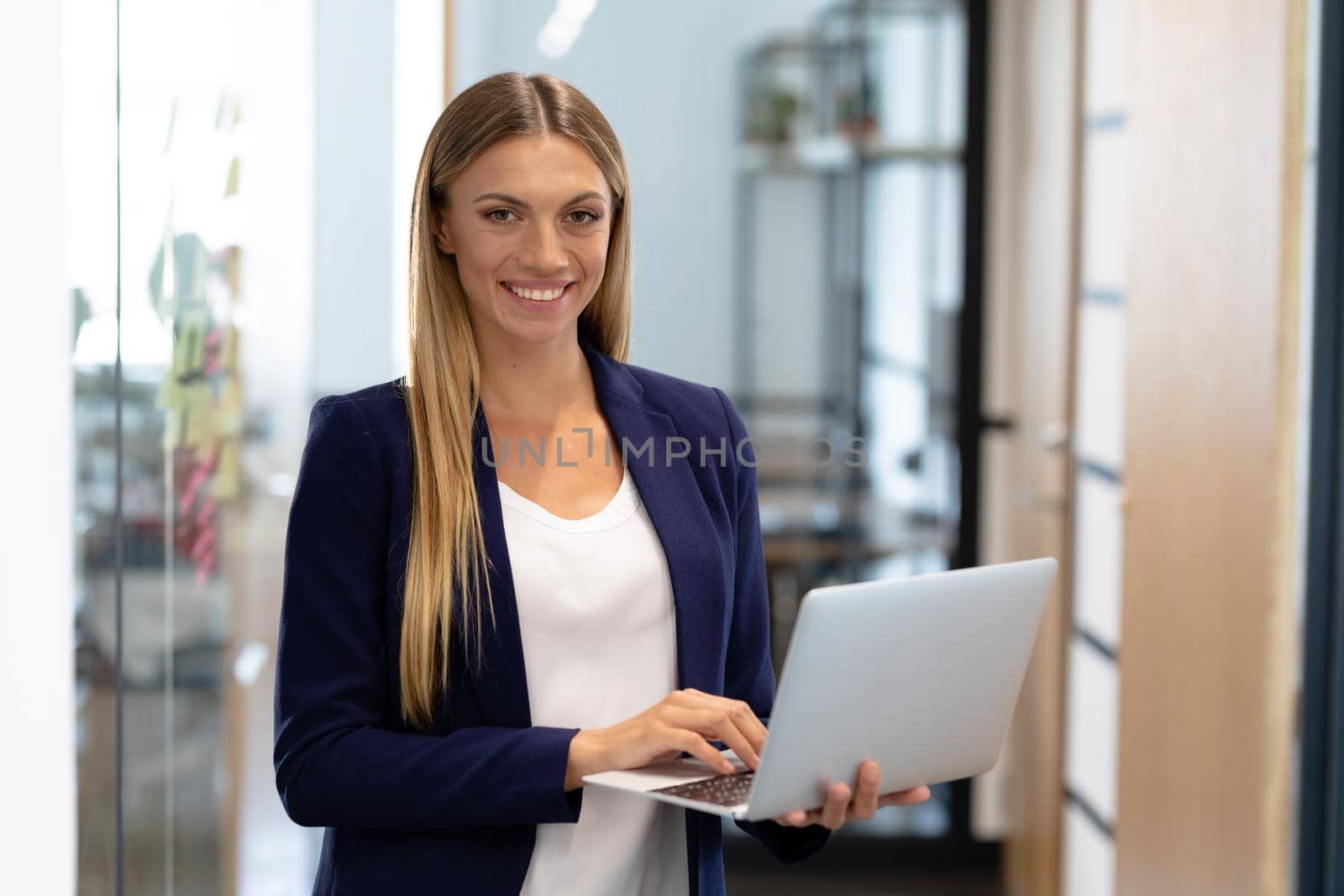 Happy caucasian businesswoman standing in corridor using laptop looking to camera by Wavebreakmedia