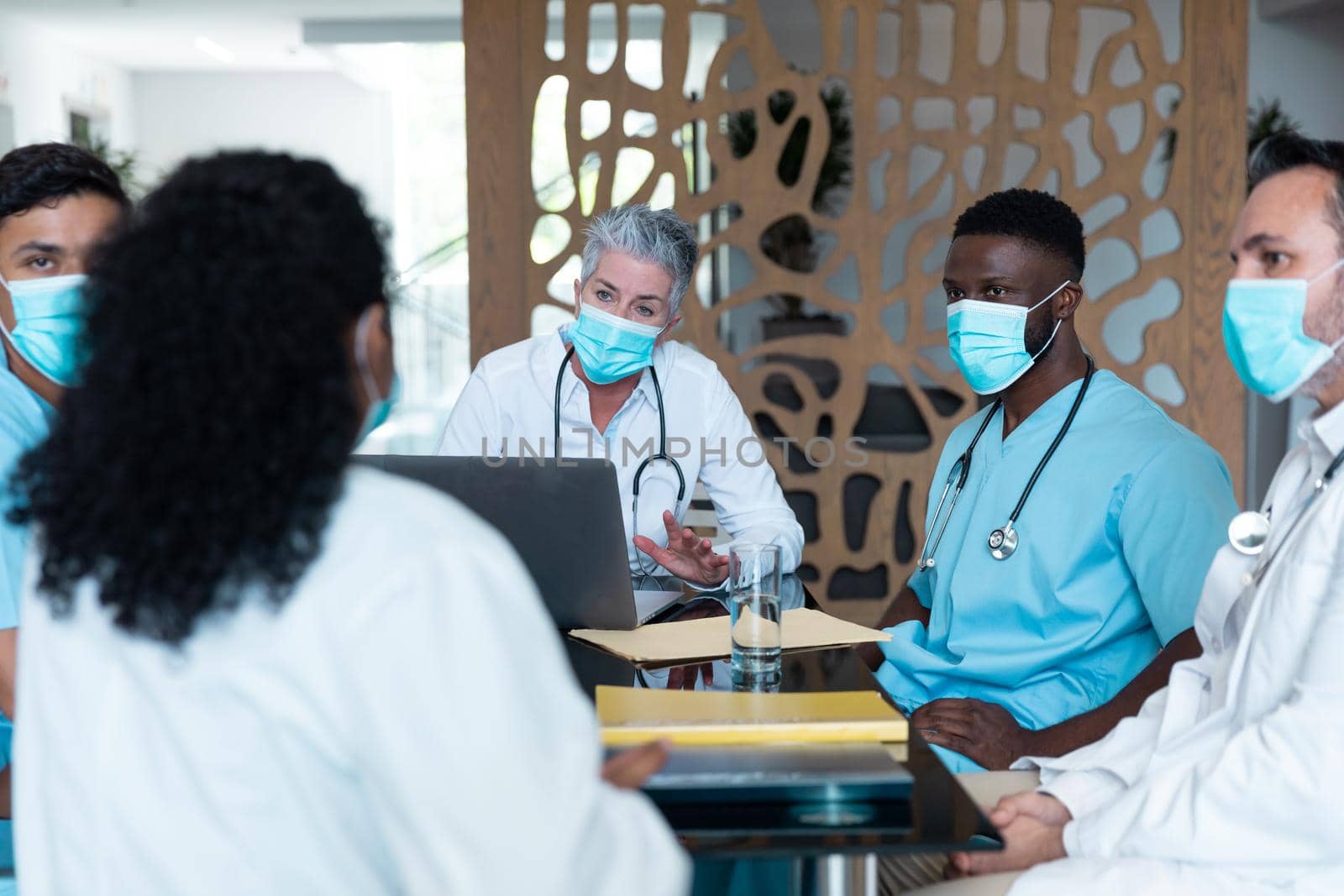 Caucasian female senior doctor and diverse hospital colleagues wearing face masks sitting at meeting. medicine, health and healthcare services during covid 19 coronavirus pandemic.