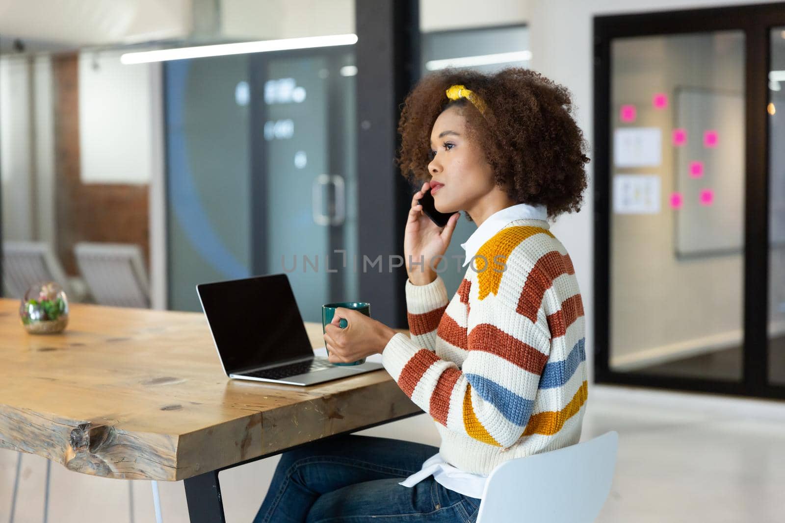 Portrait of mixed race businesswoman sitting at desk with laptop and talking on smartphone by Wavebreakmedia