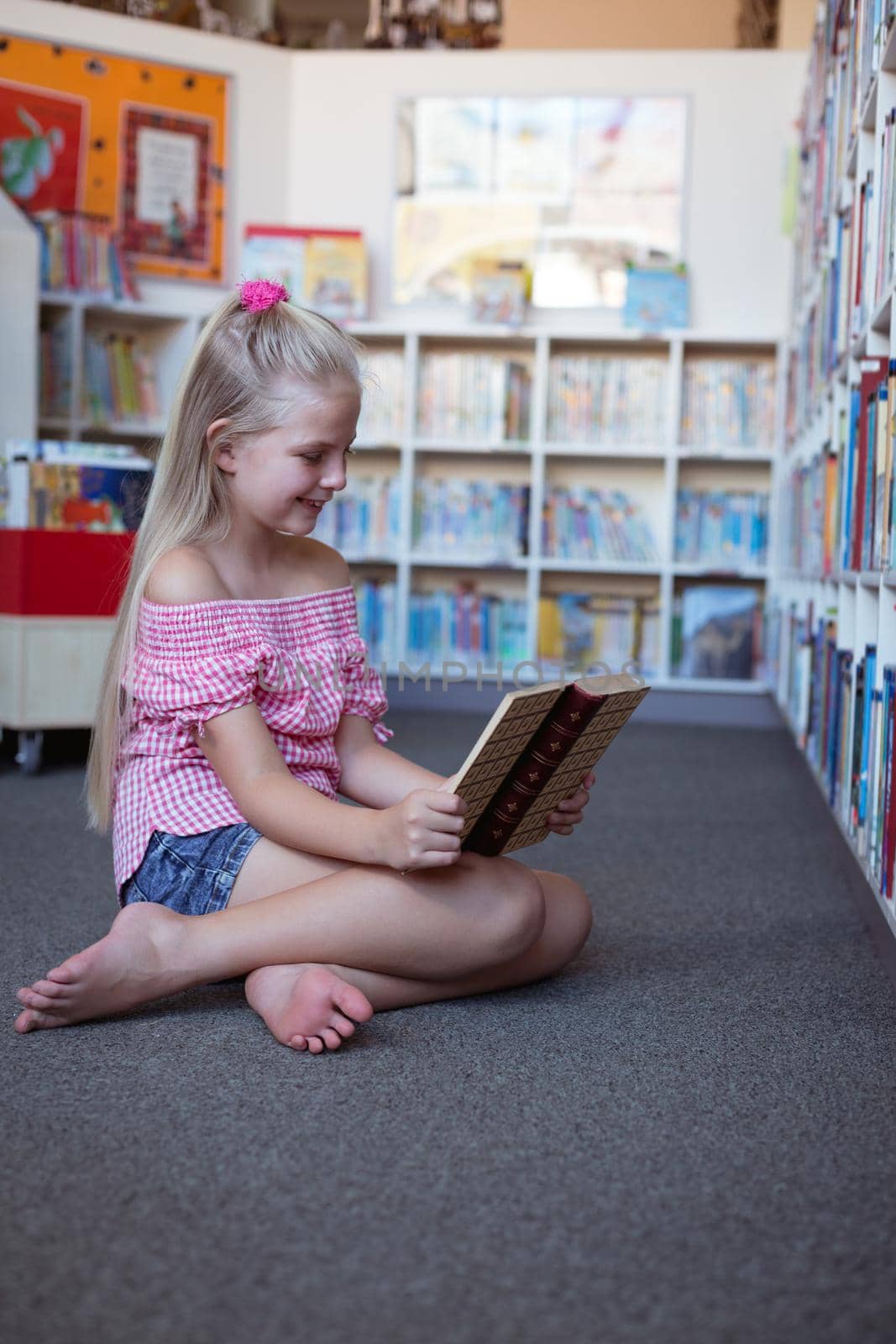 Smiling caucasian schoolgirl sitting on floor reading book in school library by Wavebreakmedia