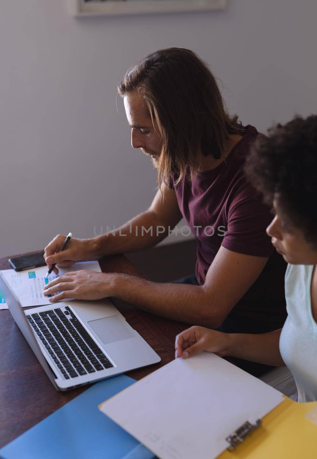 Diverse couple sitting at table working from home, using laptop and checking documents by Wavebreakmedia