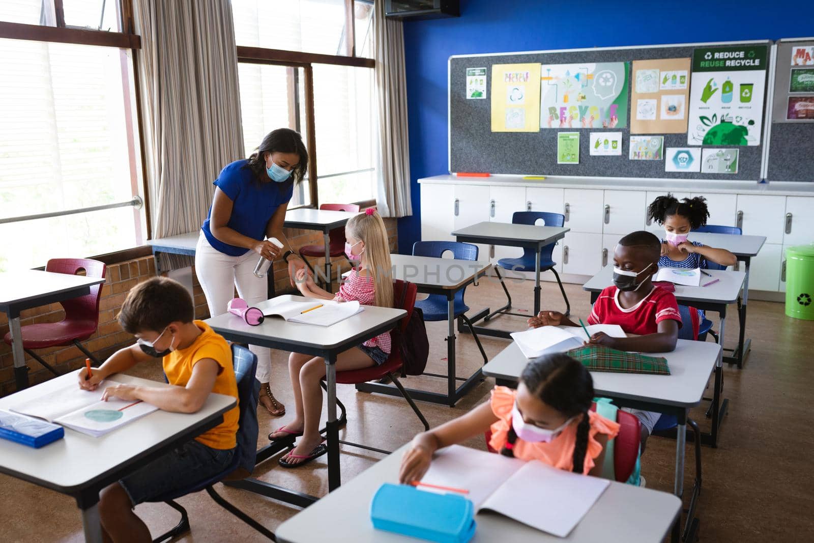 African american female teacher spraying hand sanitizer on hands of girl at elementary school. education back to school health safety during covid19 coronavirus pandemic
