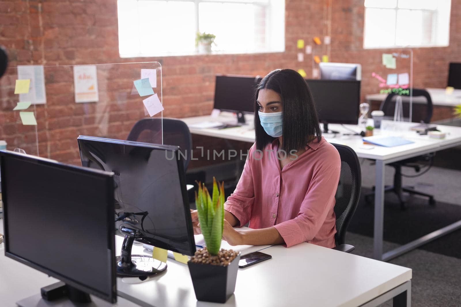 Mixed race businesswoman sitting in office in front of computer wearing face mask. independent creative design business during covid 19 coronavirus pandemic.