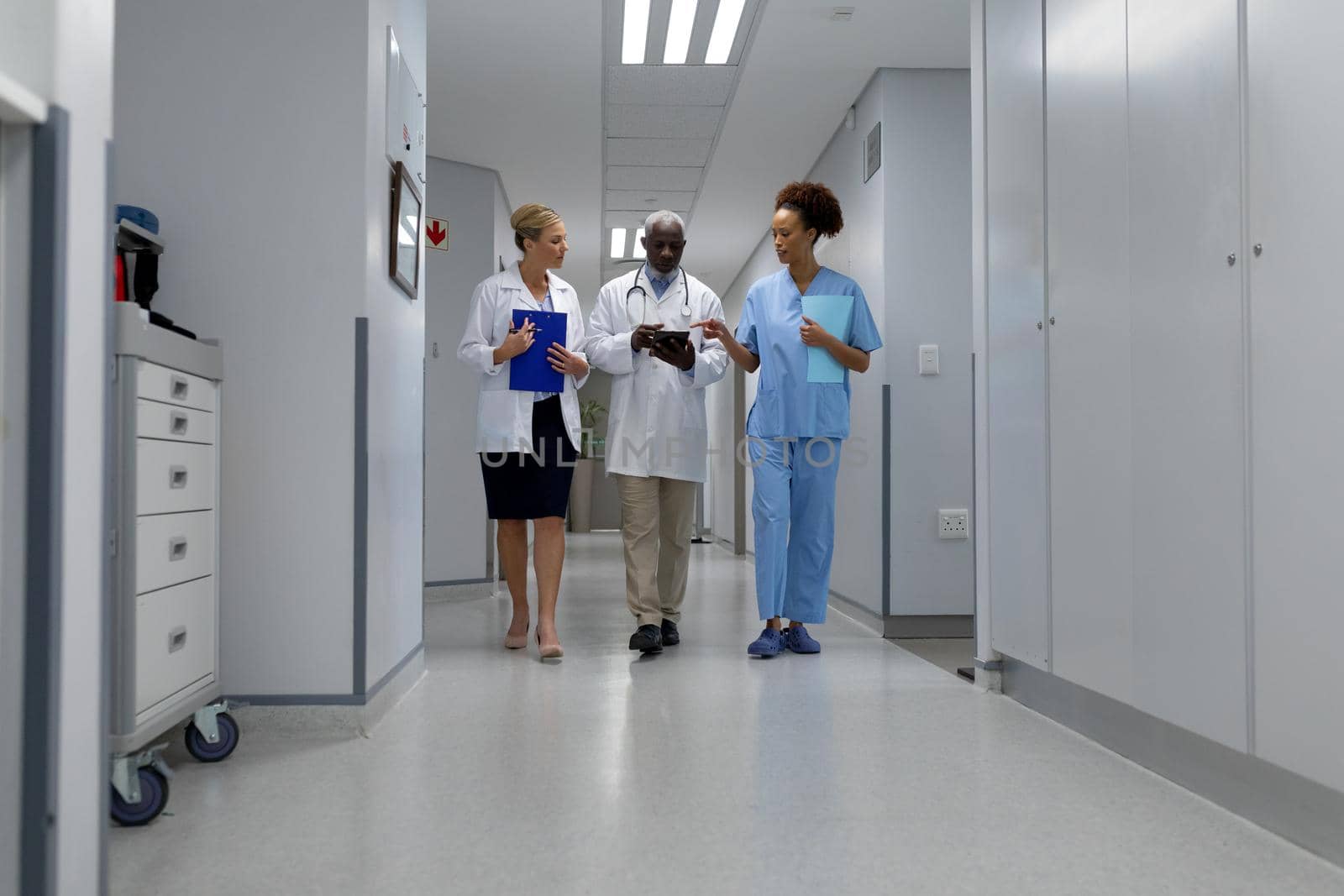 Three diverse male and female doctors walking through hospital corridor looking at digital tablet. medicine, health and healthcare services.