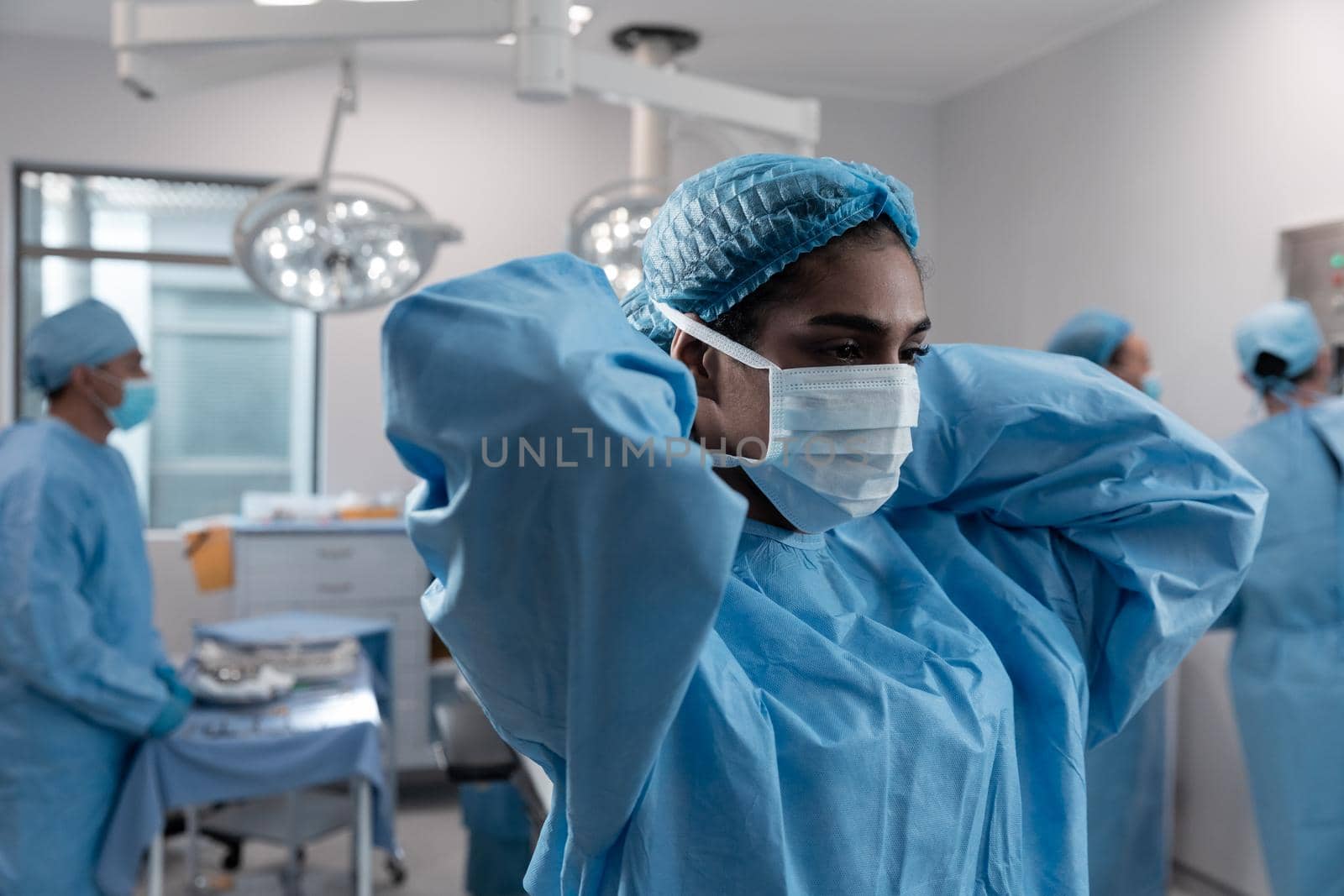 Mixed race female doctor putting on face mask standing in operating theatre. medicine, health and healthcare services during coronavirus covid 19 pandemic.