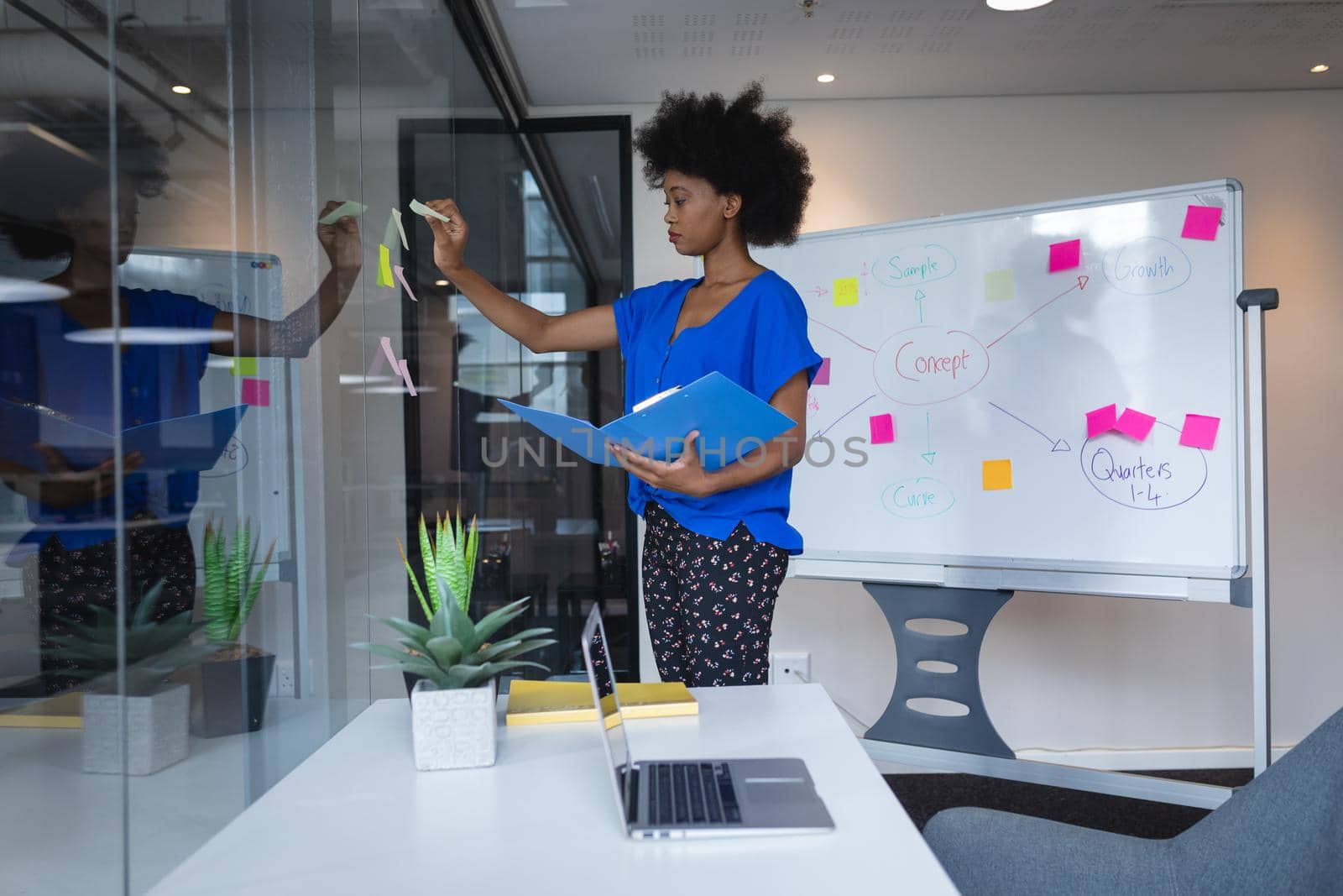 Mixed race businesswoman sticking memo notes on glass wall with whiteboard in background. independent creative design business.