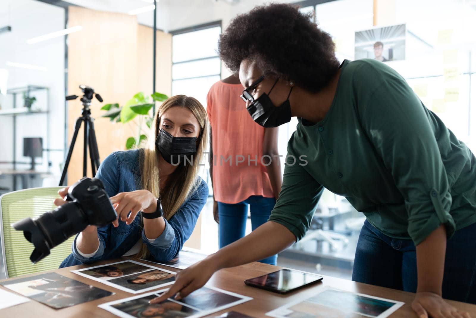 Diverse group of business colleagues wearing masks brainstorming looking at photos in meeting room. independent creative business during covid 19 coronavirus pandemic.