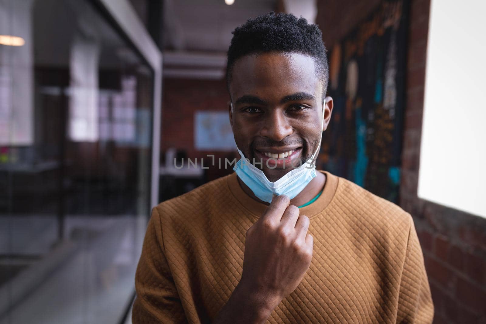 Portrait of happy african american businessman taking off face mask standing in corridor in office. independent creative design business during covid 19 coronavirus pandemic.