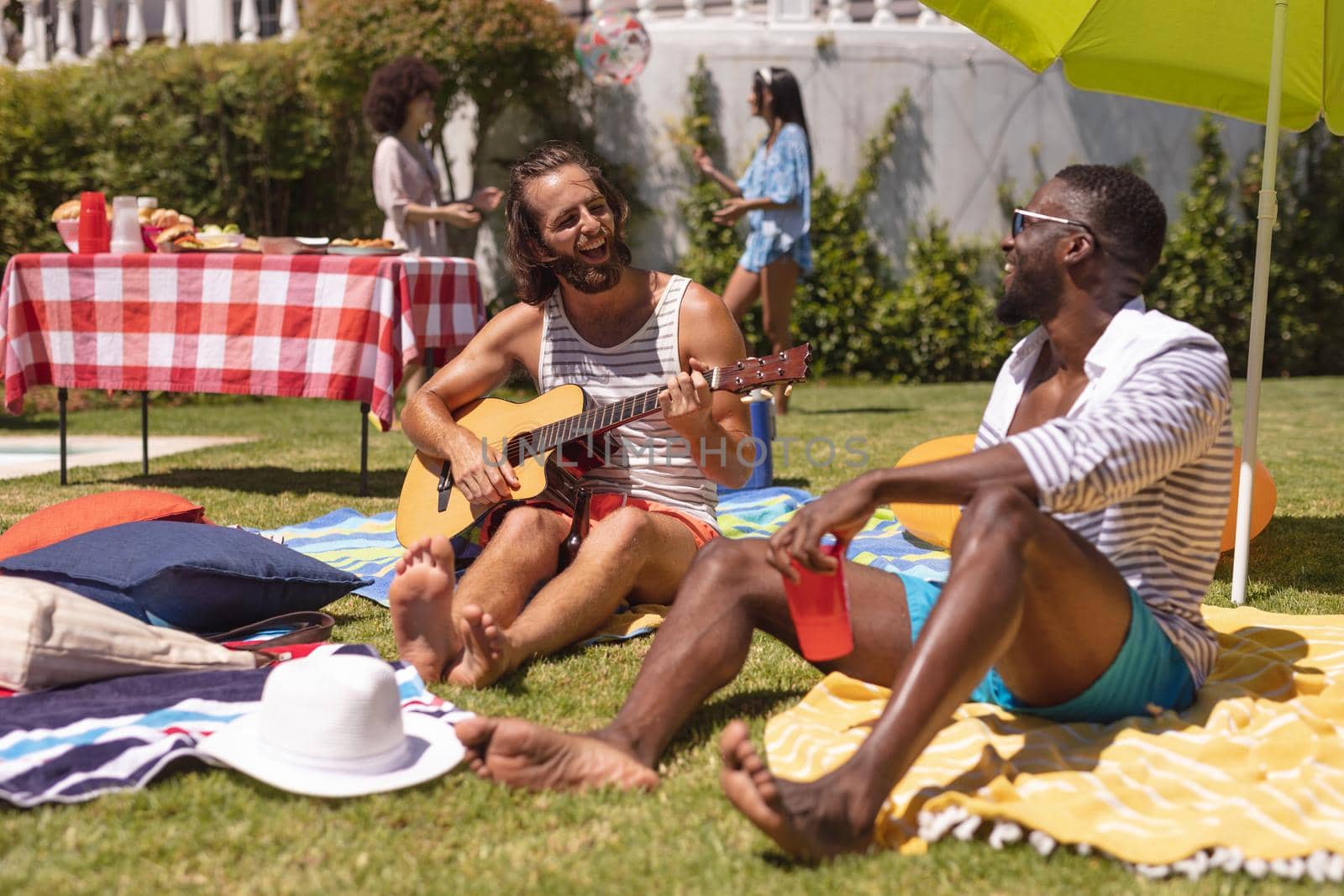 Two diverse male friends playing guitar and smiling at a pool party. Hanging out and relaxing outdoors in summer.