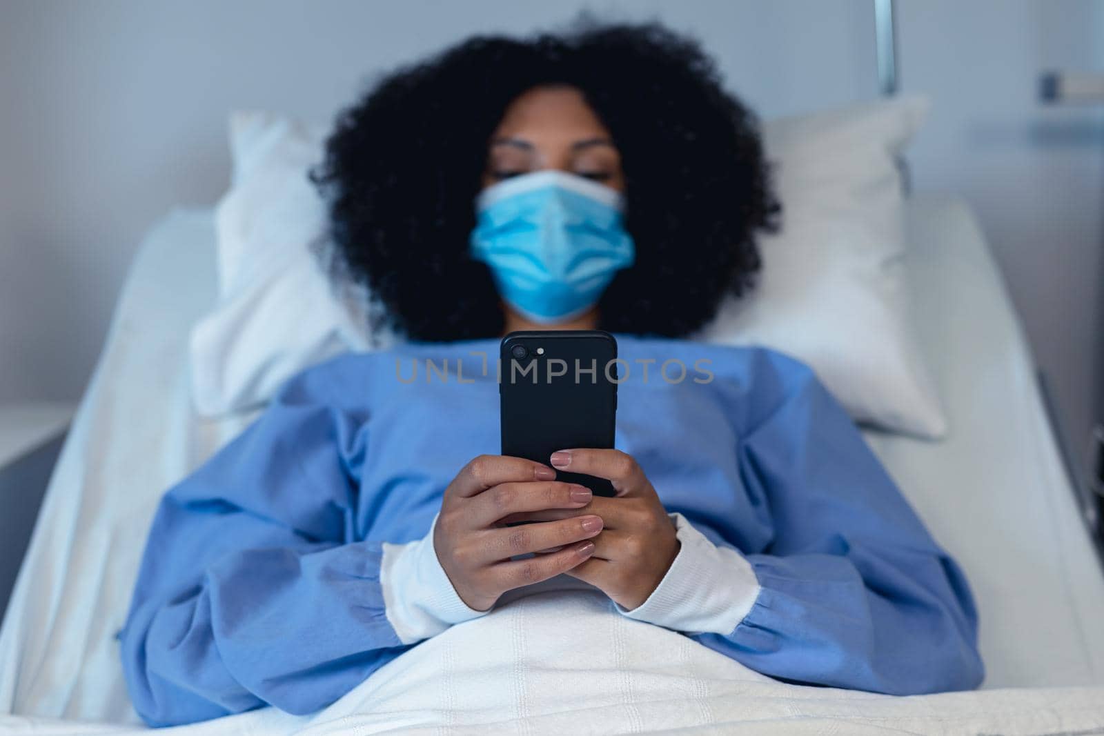 African american female patient lying in hospital bed wearing face mask using smartphone. medicine and health services during coronavirus covid 19 pandemic.