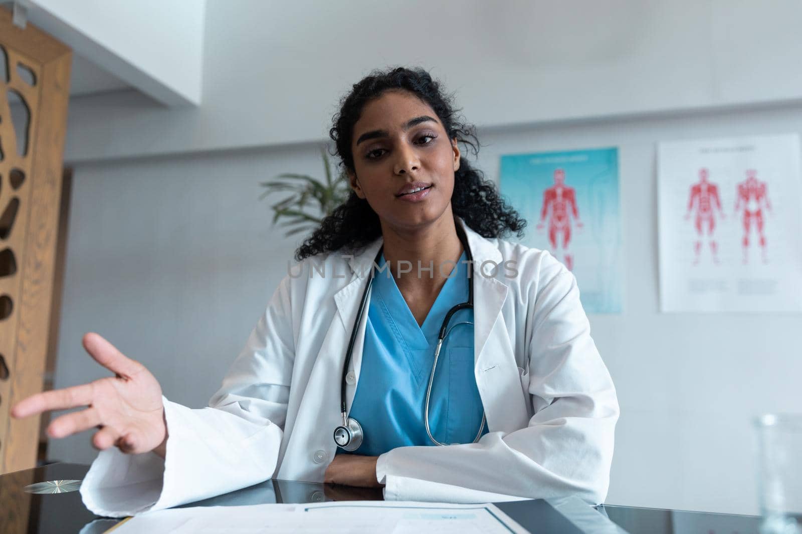 Mixed race female doctor at desk talking and gesturing during video call consultation by Wavebreakmedia