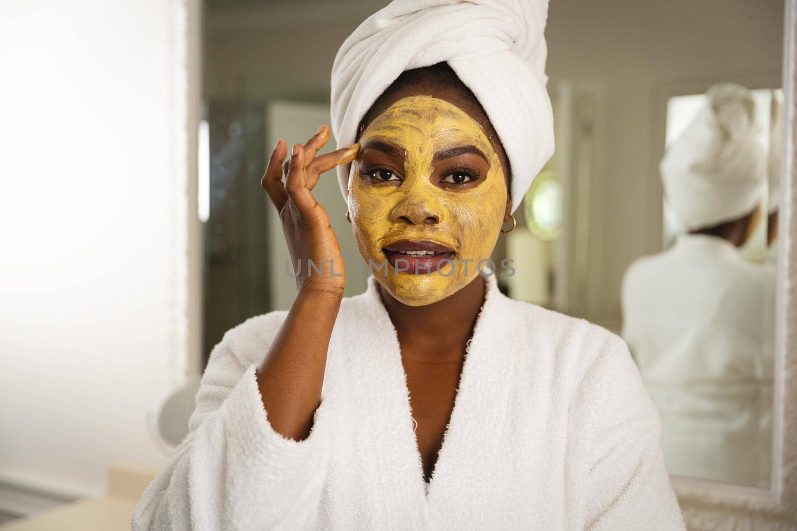 Happy african american woman in bathroom with towel on head, looking in mirror applying face mask. health, beauty and wellbeing, spending quality time at home.