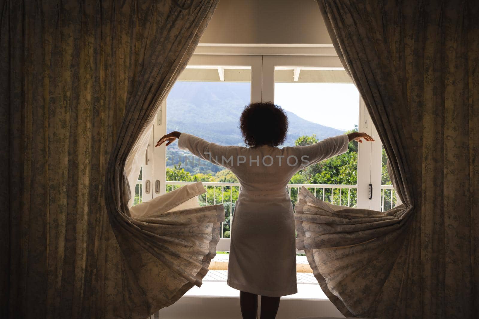African american woman standing in front of window with sunny mountain view drawing curtains by Wavebreakmedia