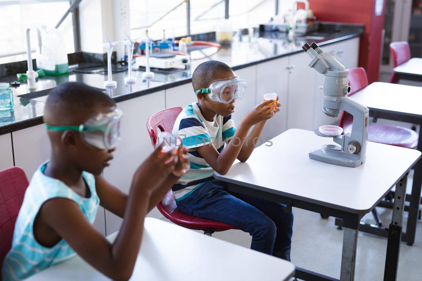 Two african american boys wearing protective glasses holding a beaker in science class at laboratory. school and education concept