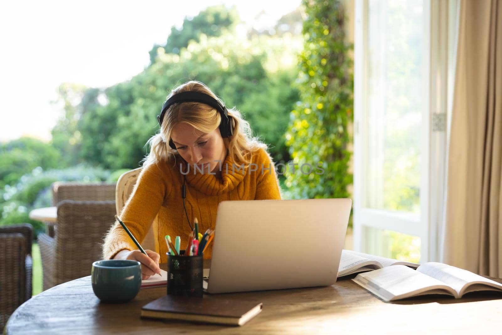 Caucasian woman working in living room at home, wearing headphones and using laptop, making notes. flexible working from home with technology.