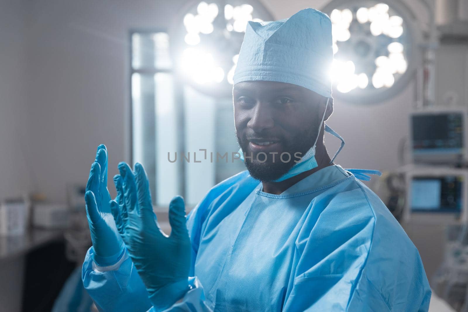 Smiling african american male surgeon with face mask in protective clothing in operating theatre. medicine, health and healthcare services during covid 19 coronavirus pandemic.