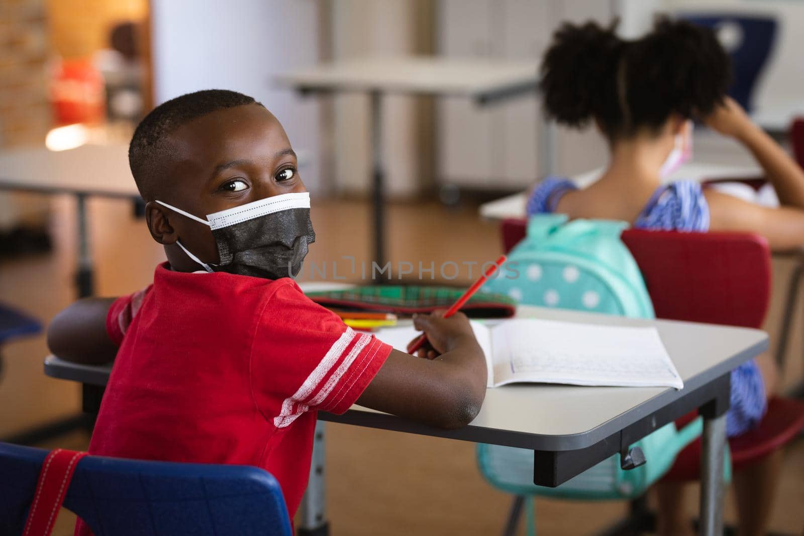 Portrait of african american boy wearing face mask sitting on his desk in class at elementary school by Wavebreakmedia