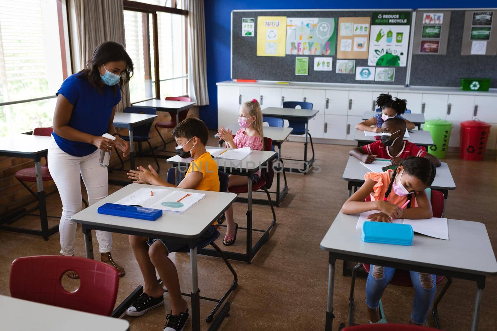 African american female teacher spraying hand sanitizer on hands of boy at elementary school. education back to school health safety during covid19 coronavirus pandemic