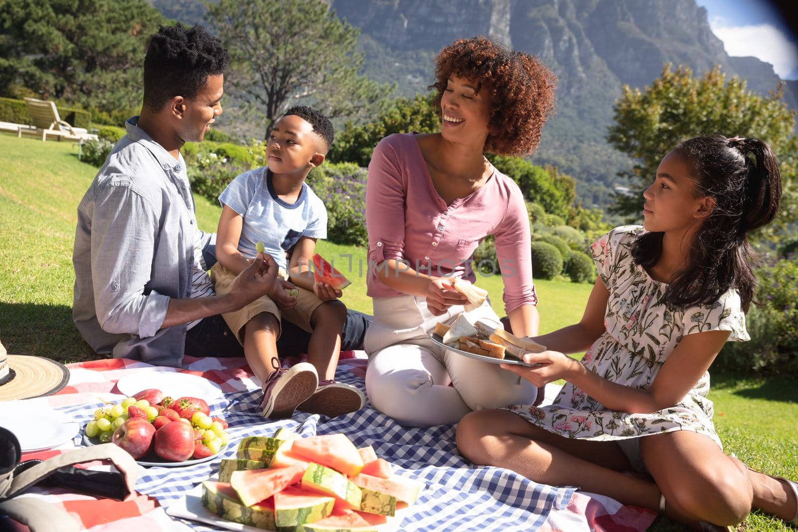 Happy african american couple with son and daughter outdoors, having picnic in sunny garden. family enjoying quality free time together.