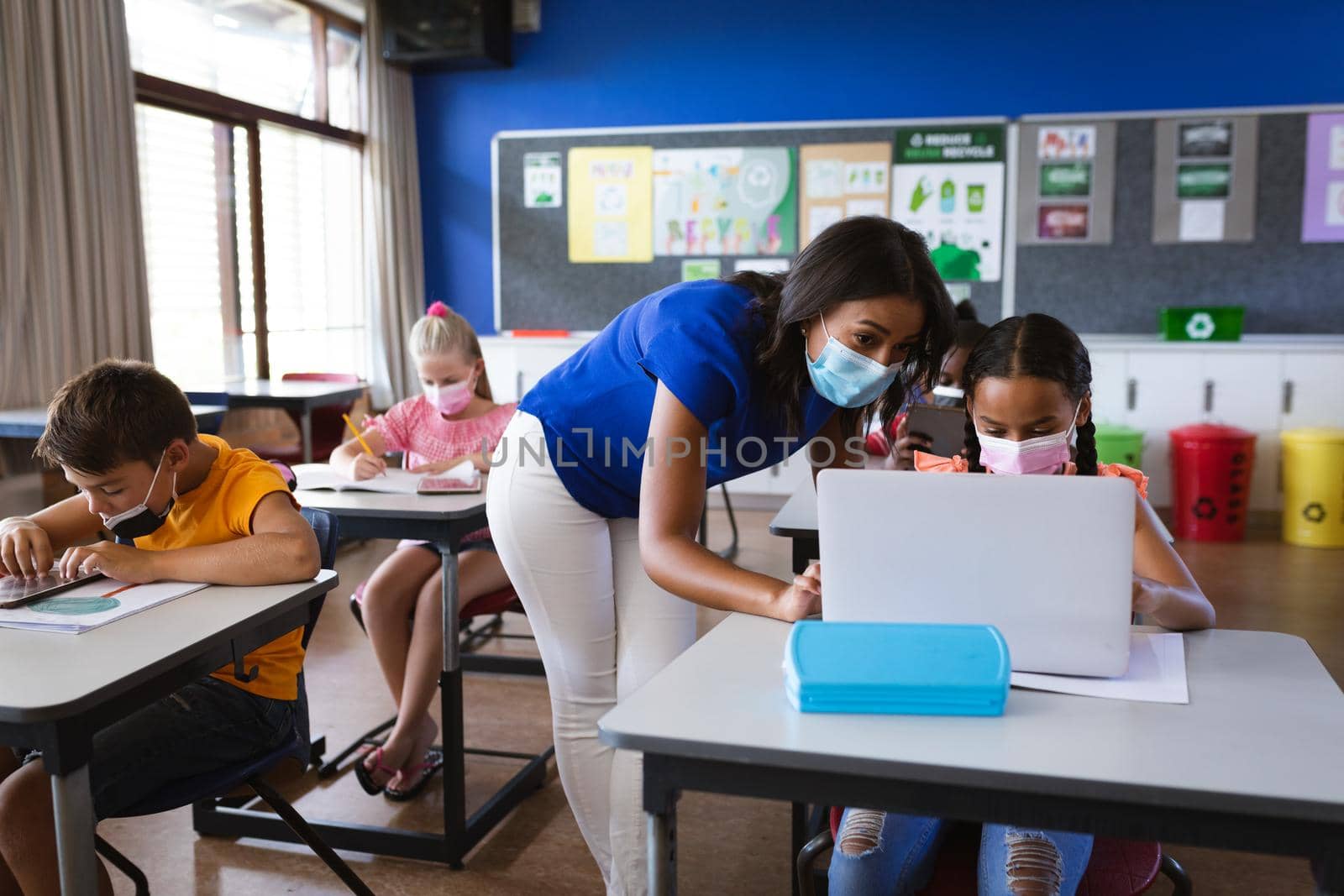 African american female teacher wearing face mask teaching a girl to use laptop at elementary school. back to school and education concept