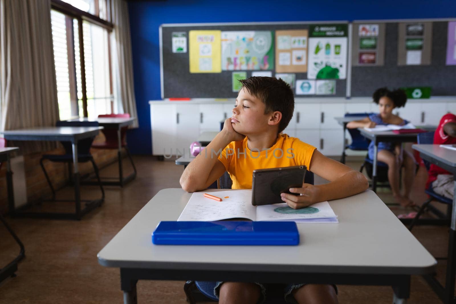 Caucasian boy holding digital tablet while sitting on the desk in the class at elementary school. school and education concept