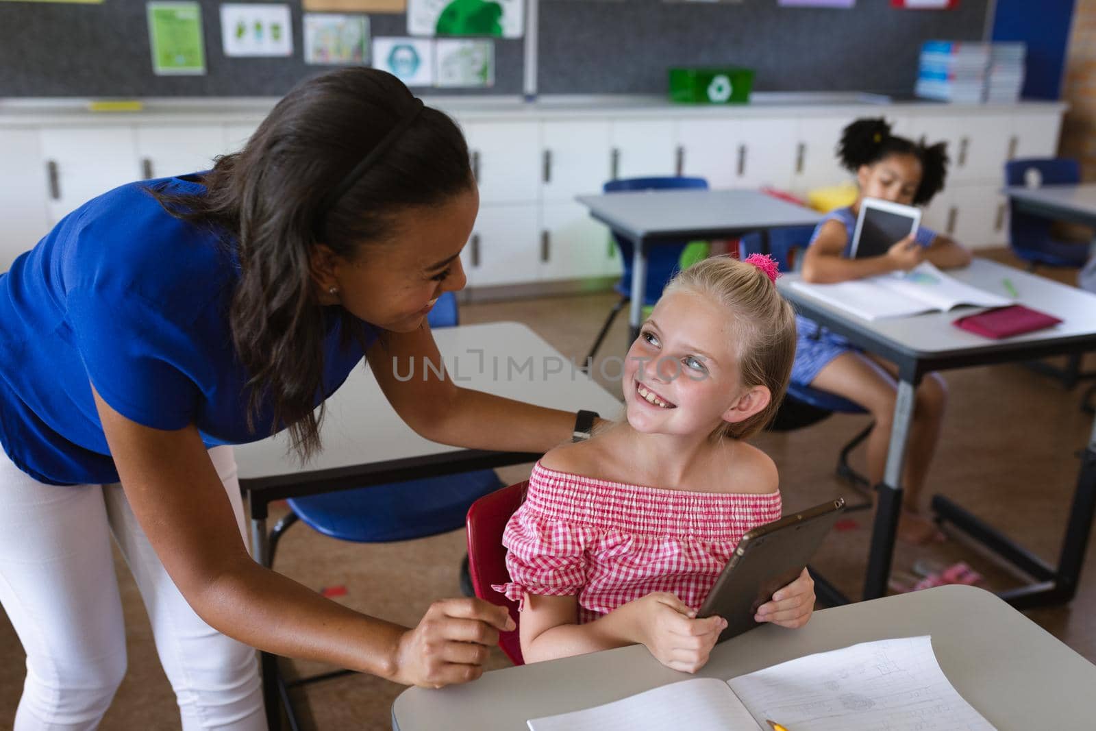 African american female teacher teaching caucasian girl to use digital tablet at elementary school. school and education concept