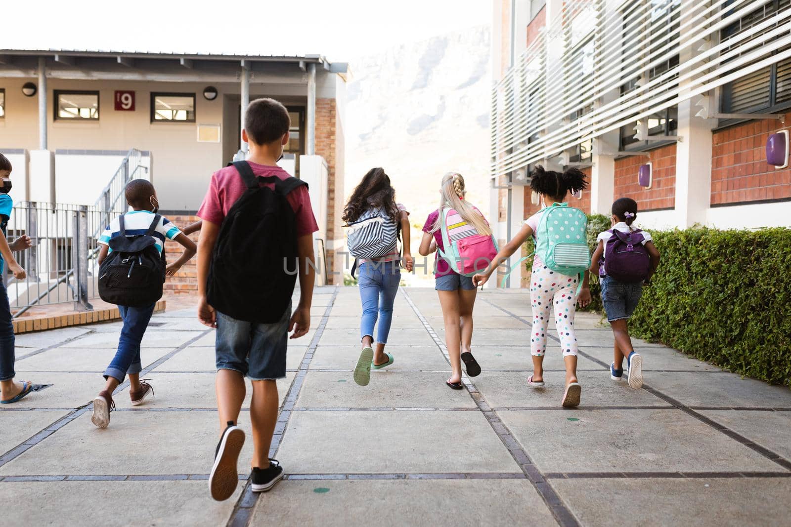 Rear view of group of diverse students with backpacks running at elementary school. education back to school health safety during covid19 coronavirus pandemic.