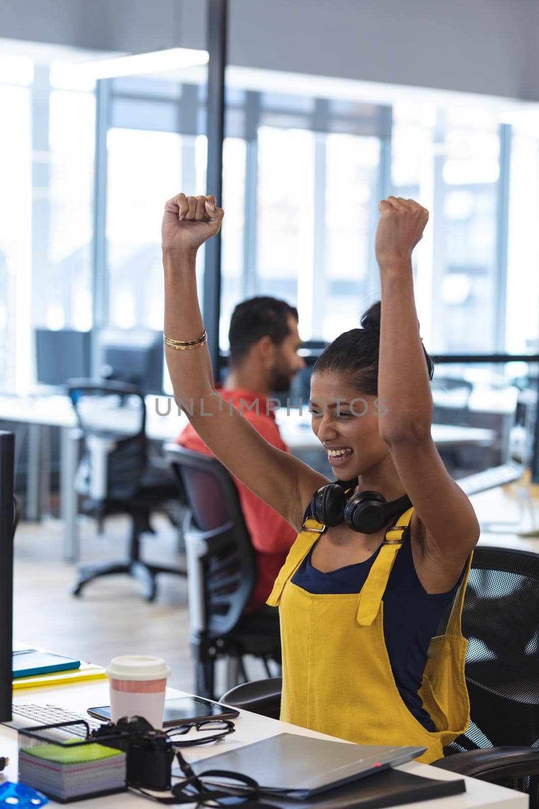 Mixed race female creative sitting at desk celebrating raising her arms and smiling. modern office of a creative design business.