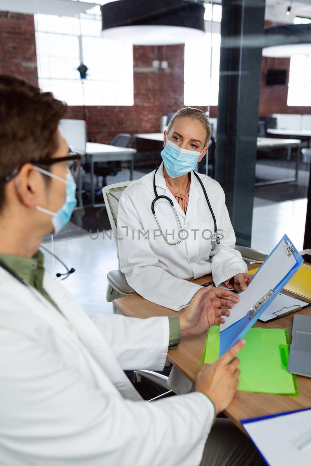 Diverse male and female doctor wearing face masks sitting in hospital office discussing paperwork. medical and healthcare services during coronavirus covid 19 pandemic.