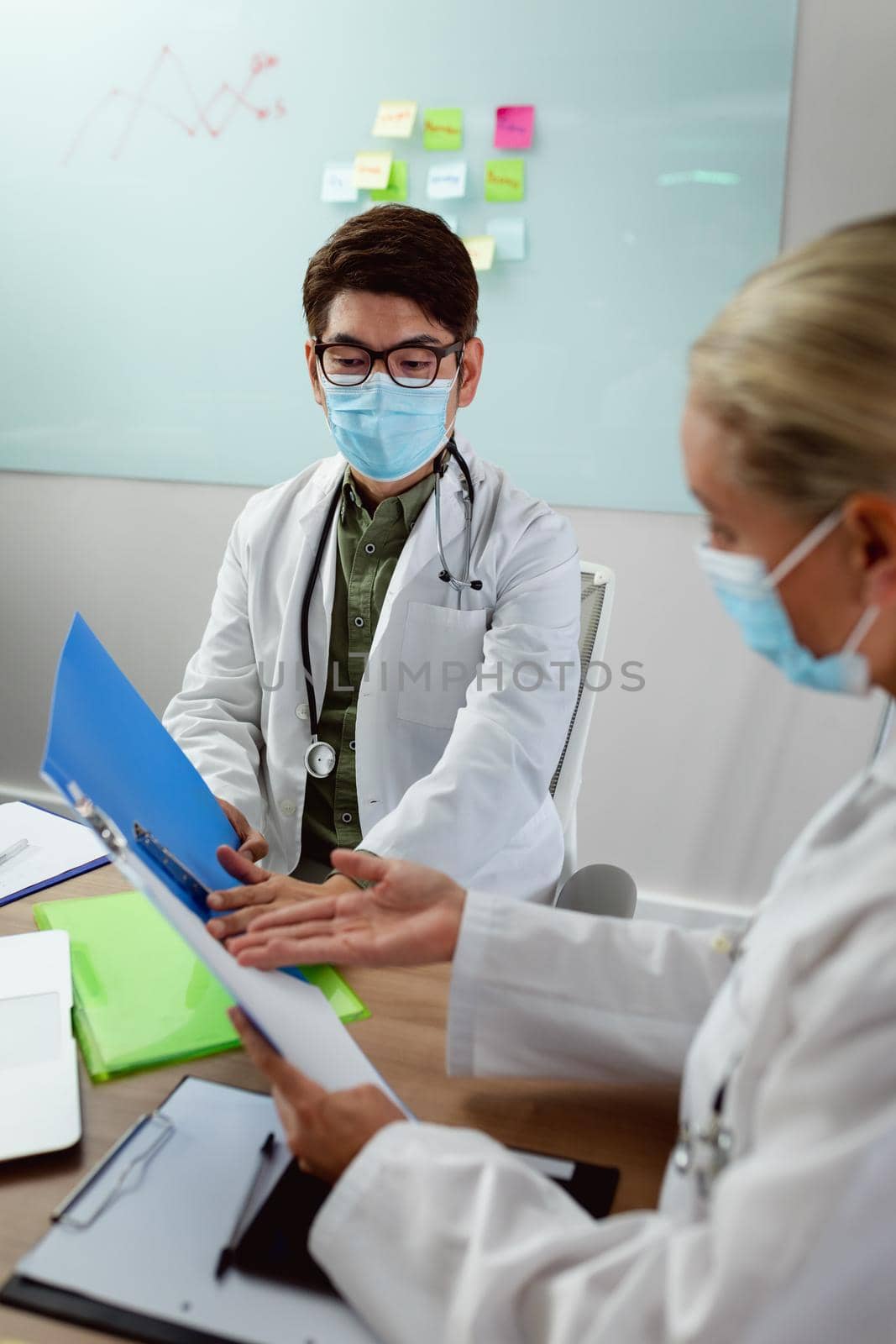 Diverse male and female doctor wearing face masks sitting in hospital office discussing paperwork by Wavebreakmedia