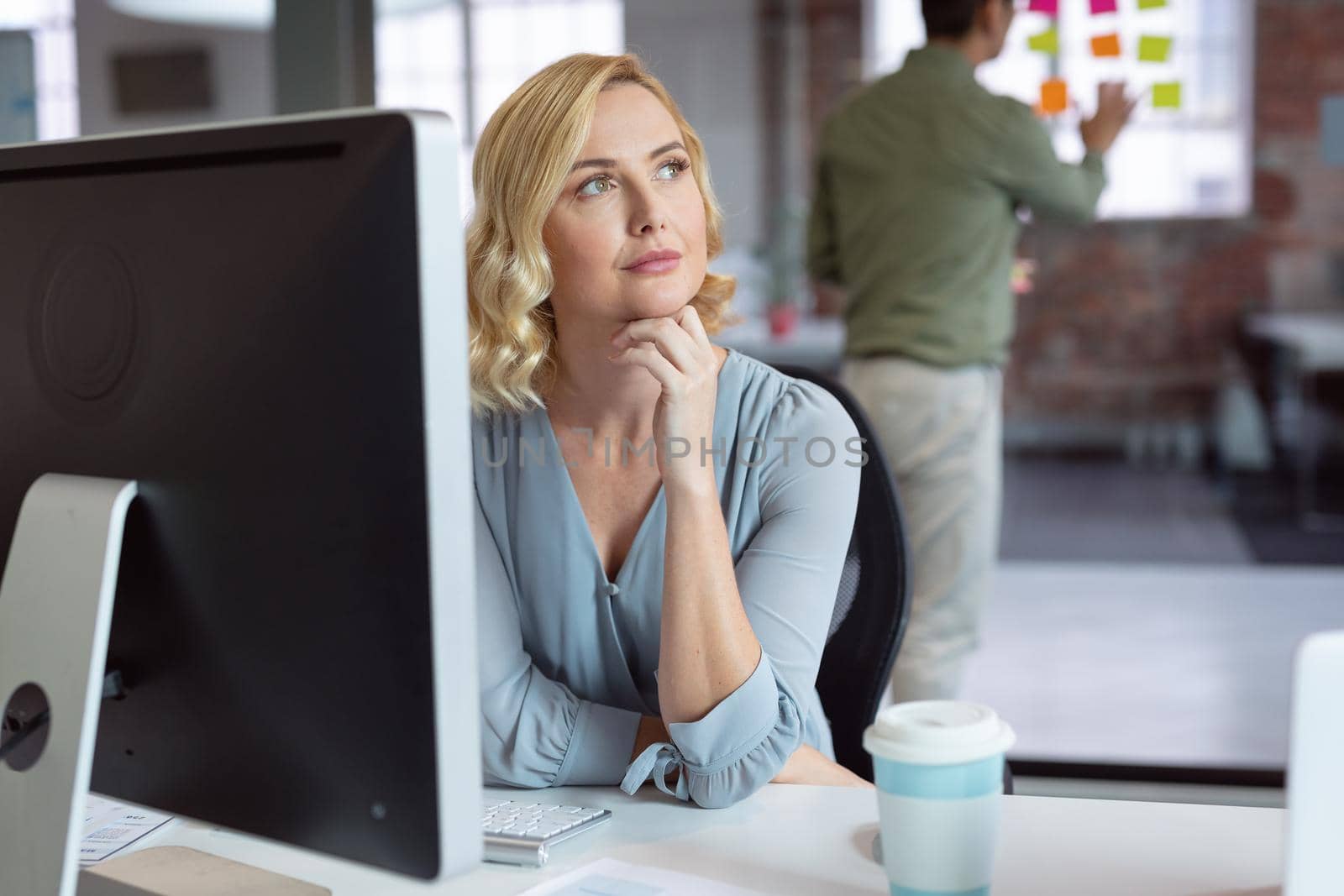 Thoughtful caucasian businesswoman sitting at desk using computer with male colleague in background. working in business at a modern office.
