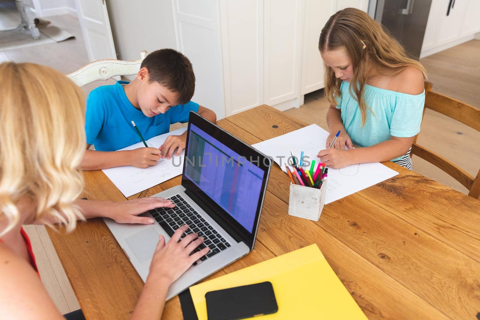 Caucasian mother using laptop and her daughter and son doing homework in kitchen. family domestic life, spending time working together at home.