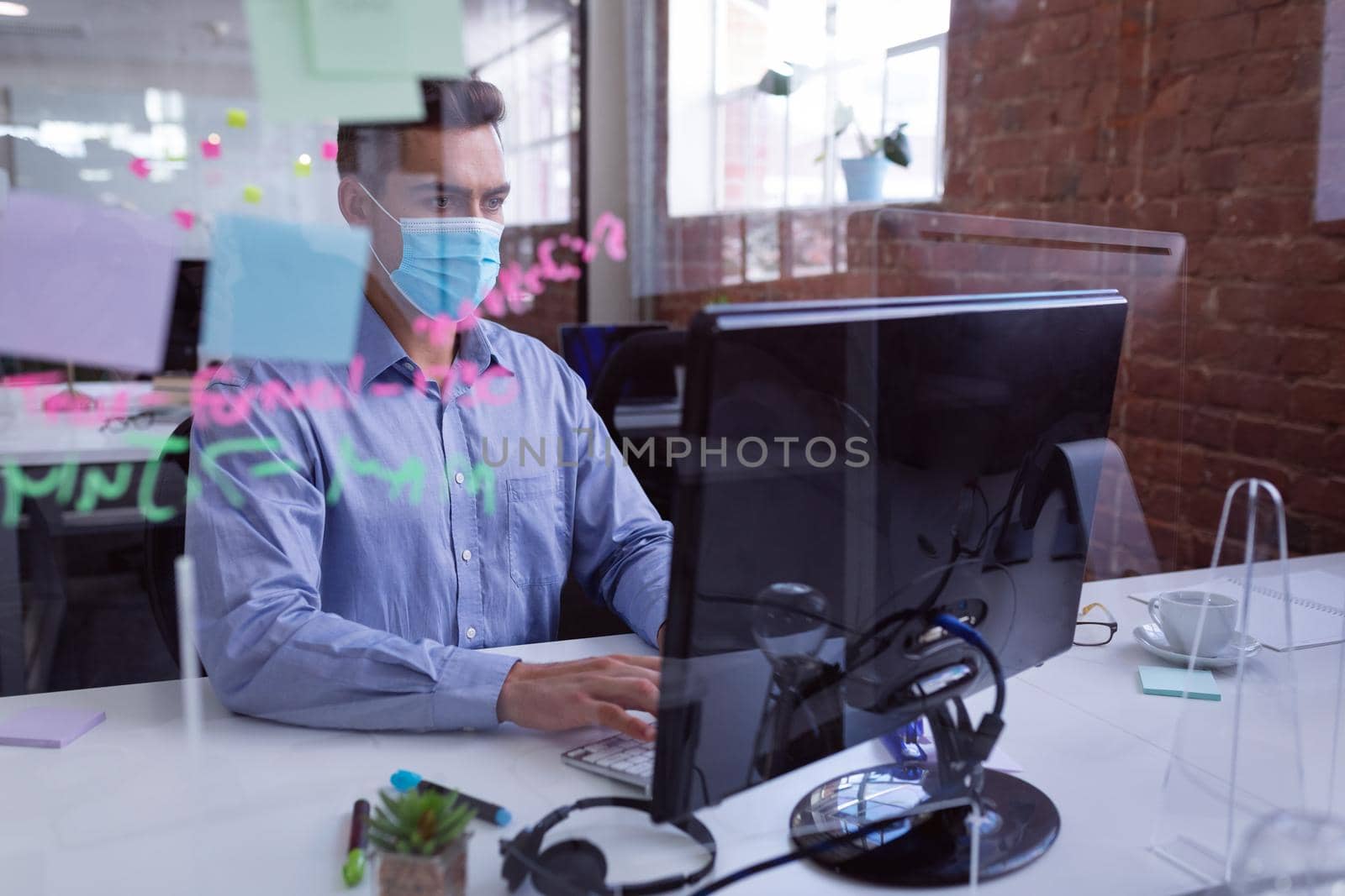 Caucasian businessman in face mask sitting at desk with hygiene screen using computer by Wavebreakmedia