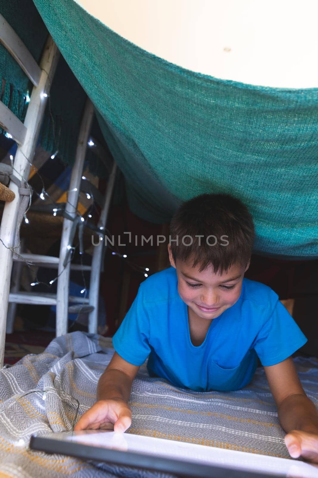 Caucasian boy smiling, lying in blanket fort using tablet at home. childhood with technology, spending free time at home.