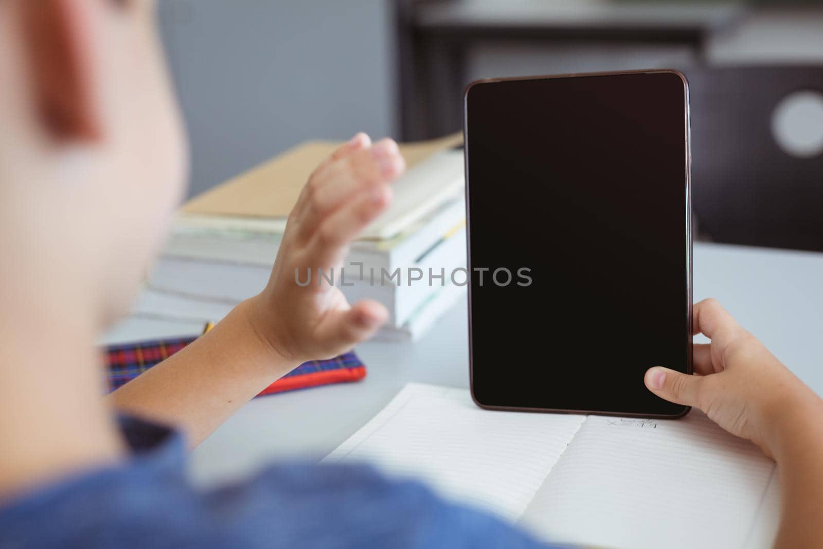 Caucasian schoolboy in classroom sitting at desk using tablet and waving, with copy space on screen by Wavebreakmedia