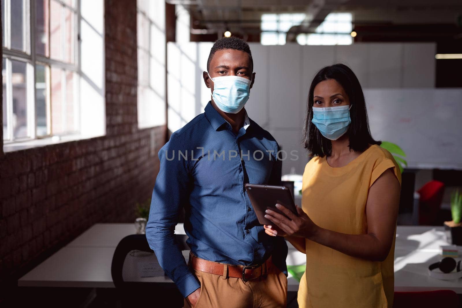 Portrait of diverse male and female colleague wearing face masks standing in office using tablet. working in business at a modern office during coronavirus covid 19 pandemic.
