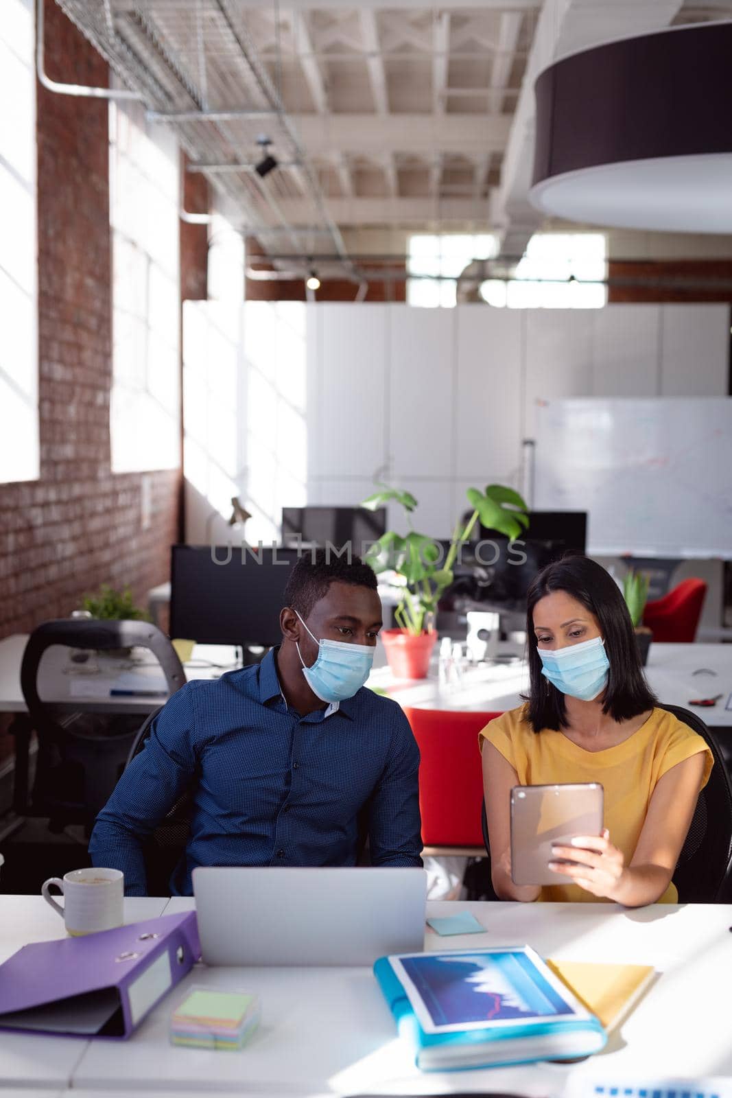 Diverse male and female colleague in face masks sitting at desk discussing, using laptop and tablet by Wavebreakmedia