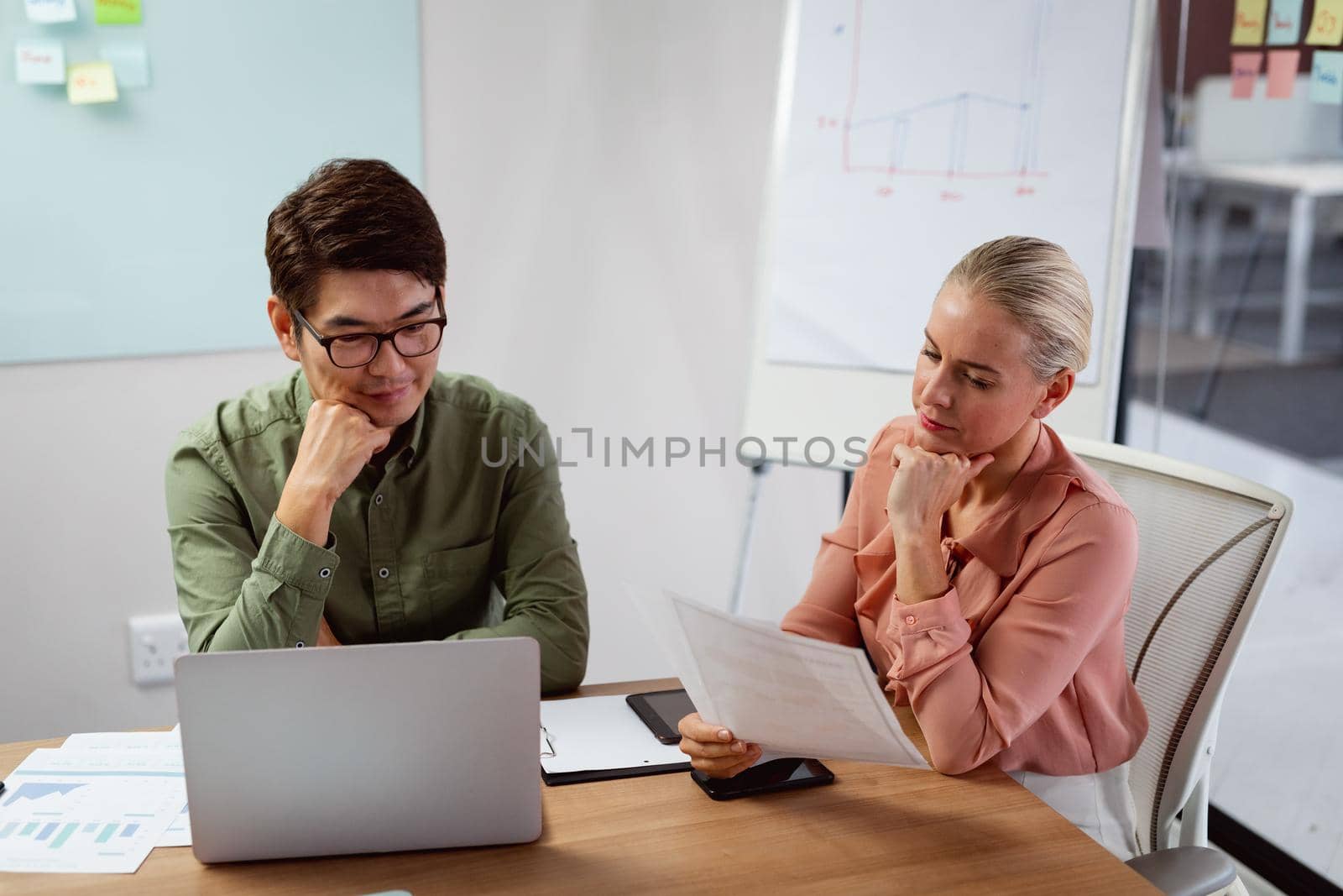 Diverse male and female colleague sitting at table with laptop and paperwork discussing by Wavebreakmedia