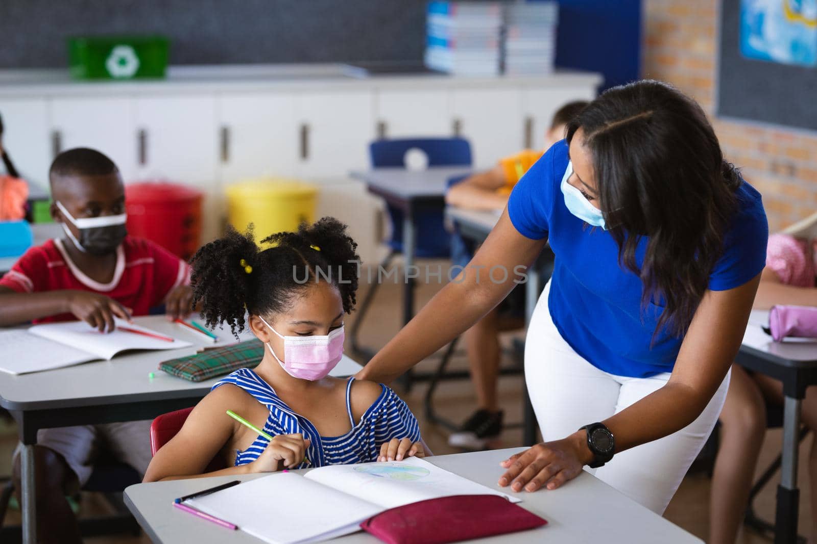 African american female teacher wearing face mask teaching african american girl in class at school by Wavebreakmedia