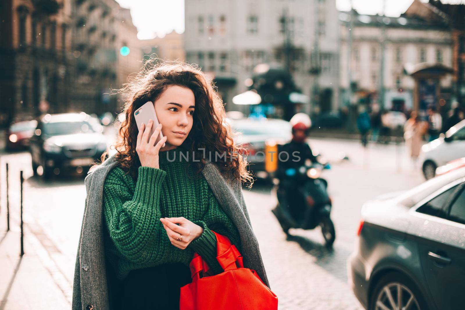 A woman on the street uses a mobile phone. online shopping. use of mobile applications. beautiful young woman with long curly dark hair in a casual coat, trendy green sweater and red handbag