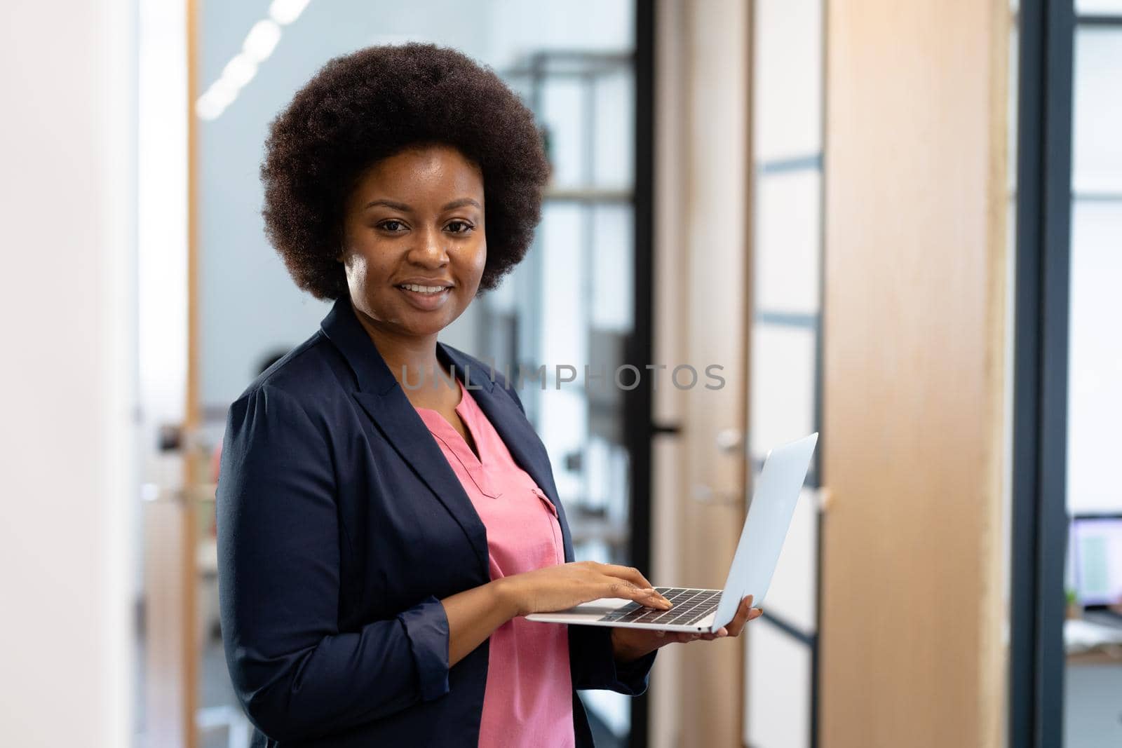 Happy african american businesswoman standing in corridor using laptop looking to camera by Wavebreakmedia