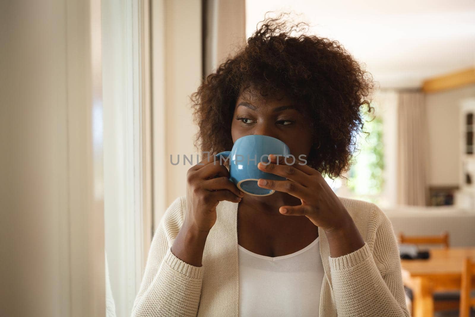 Thoughtful african american woman standing in dining room drinking cup of coffee by Wavebreakmedia