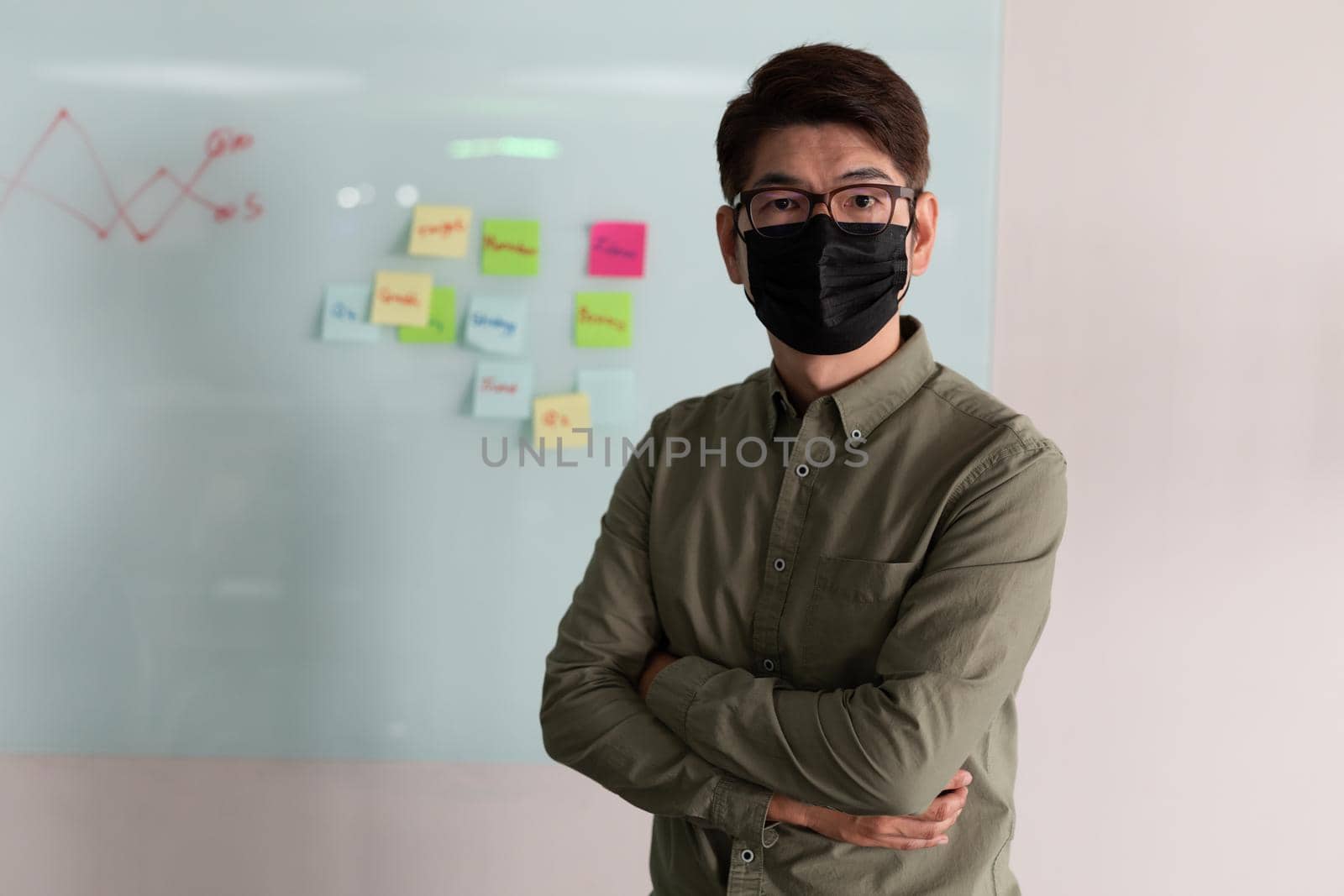 Portrait of asian businessman wearing face mask standing in meeting room looking to camera by Wavebreakmedia