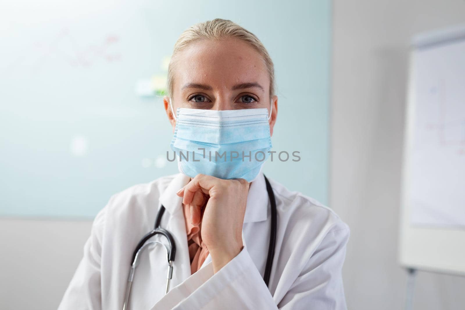 Caucasian female doctor wearing face mask sitting at desk in office listening during video call by Wavebreakmedia