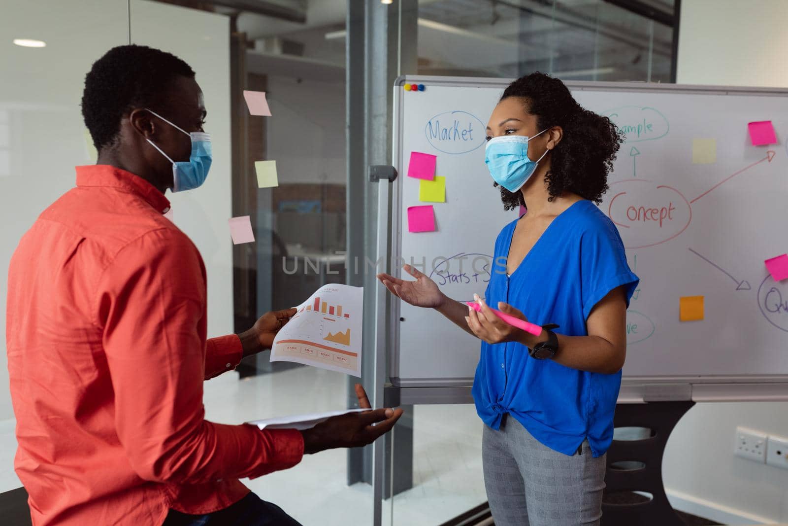 Diverse male and female colleague in face masks discussing paperwork in office. working in business at a modern office during coronavirus covid 19 pandemic.