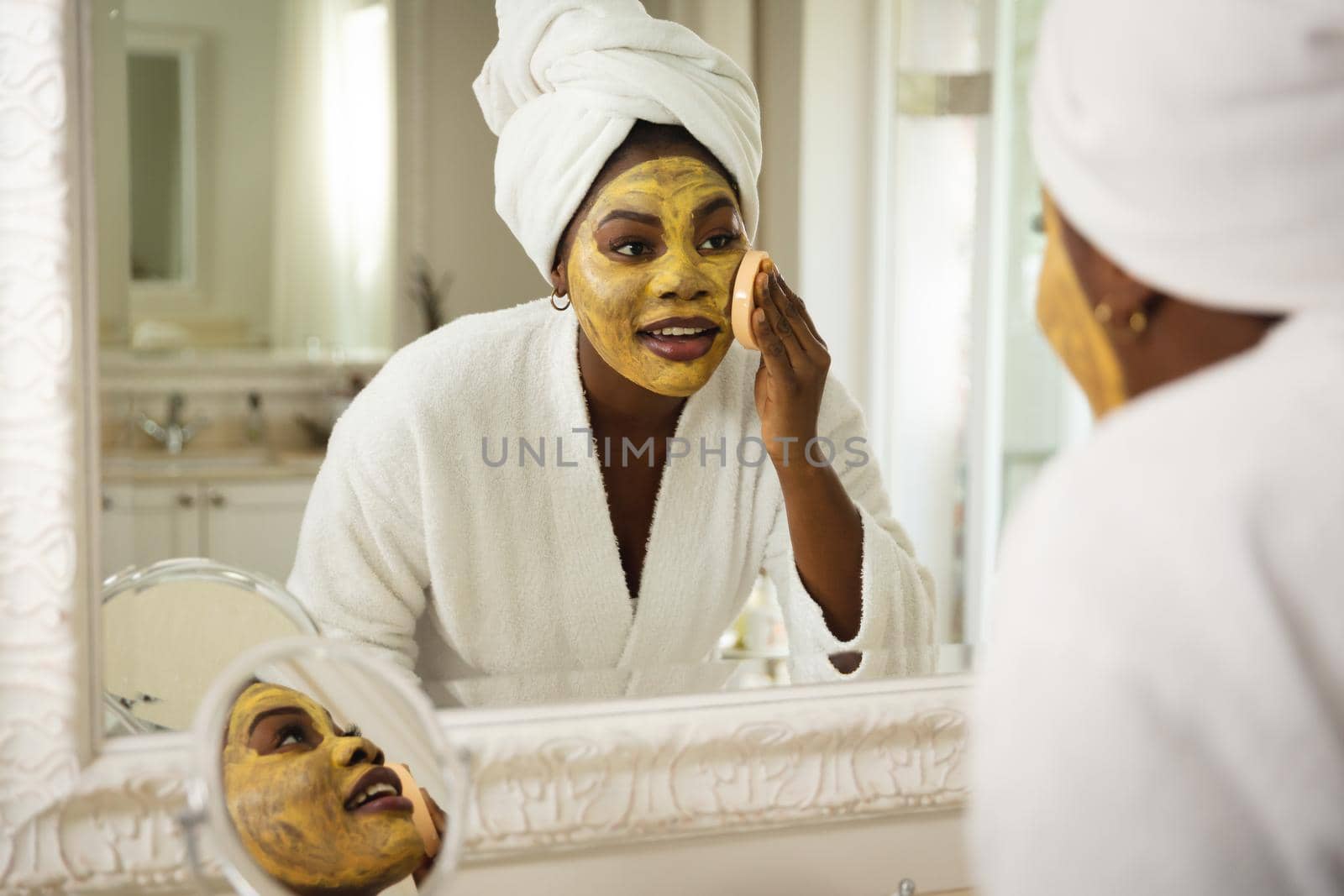 Happy african american woman in bathroom with towel on head, looking in mirror applying face mask by Wavebreakmedia