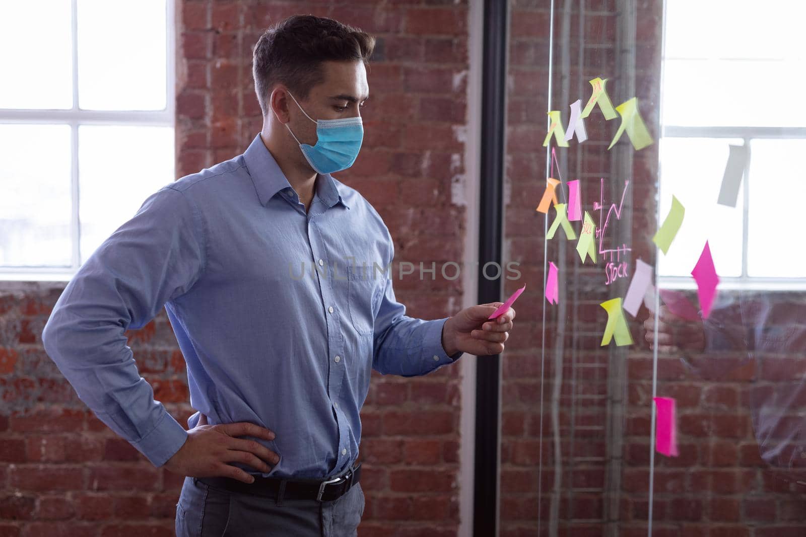 Caucasian businessman wearing face mask brainstorming, reading memo notes on wall standing in office by Wavebreakmedia