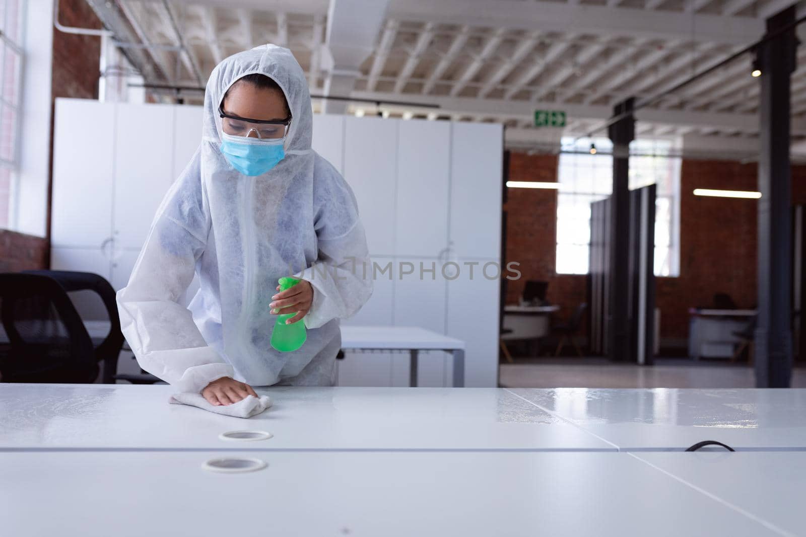 Cleaner wearing ppe suit, glasses and mask disinfecting office workspace, wiping desks by Wavebreakmedia