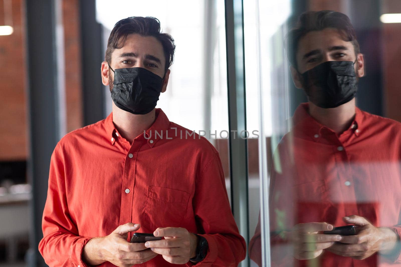 Portrait of caucasian businessman wearing face mask standing in corridor using smartphone. independent creative business during covid 19 coronavirus pandemic.