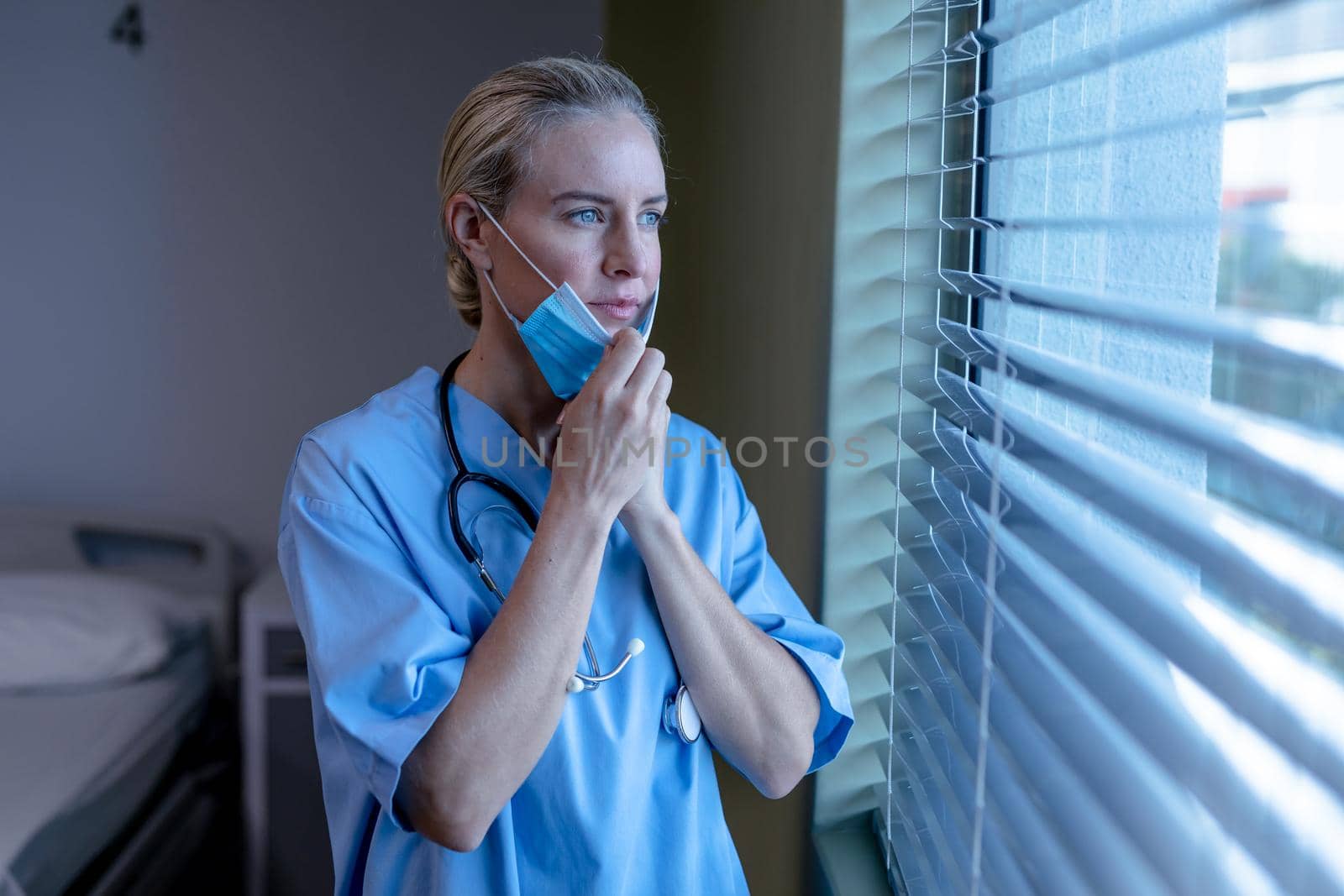Worried caucasian female doctor in hospital with face mask wearing scrubs looking out of window. medical professional at work and health services during coronavirus covid 19 pandemic.